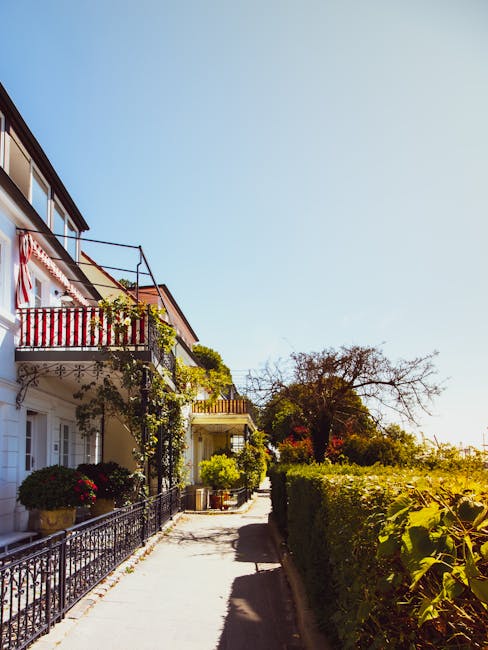 A narrow sidewalk runs parallel to a row of residential buildings on the left, featuring a mix of balconies and potted plants. The buildings display white facades with red and black railings, some of 
