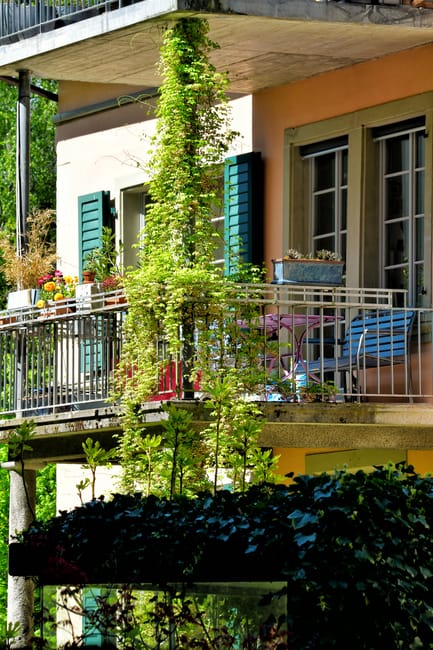 A narrow sidewalk runs parallel to a row of residential buildings on the left, featuring a mix of balconies and potted plants. The buildings display white facades with red and black railings, some of 