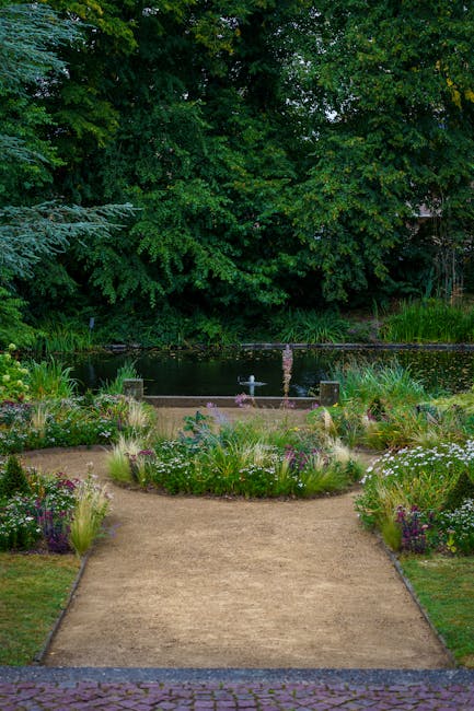 A tranquil garden scene featuring a well-maintained gravel pathway that runs through colorful flower beds, including white, purple, and green foliage, leading towards a small pond with a fountain. The